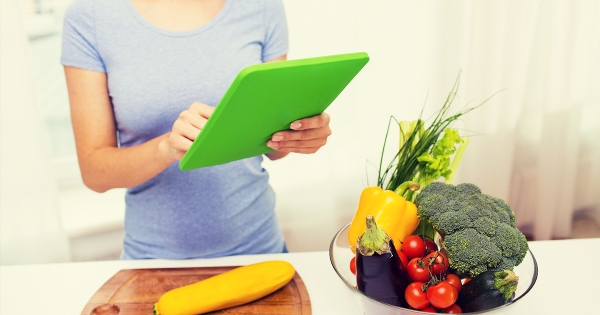 A female reviewing healthy recipes on a tablet in front of a kitchen counter topped with colorful vegetables
