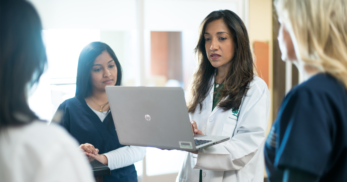 A female oncologist in a white lab coat sharing information from a laptop with female clinicians in scrubs